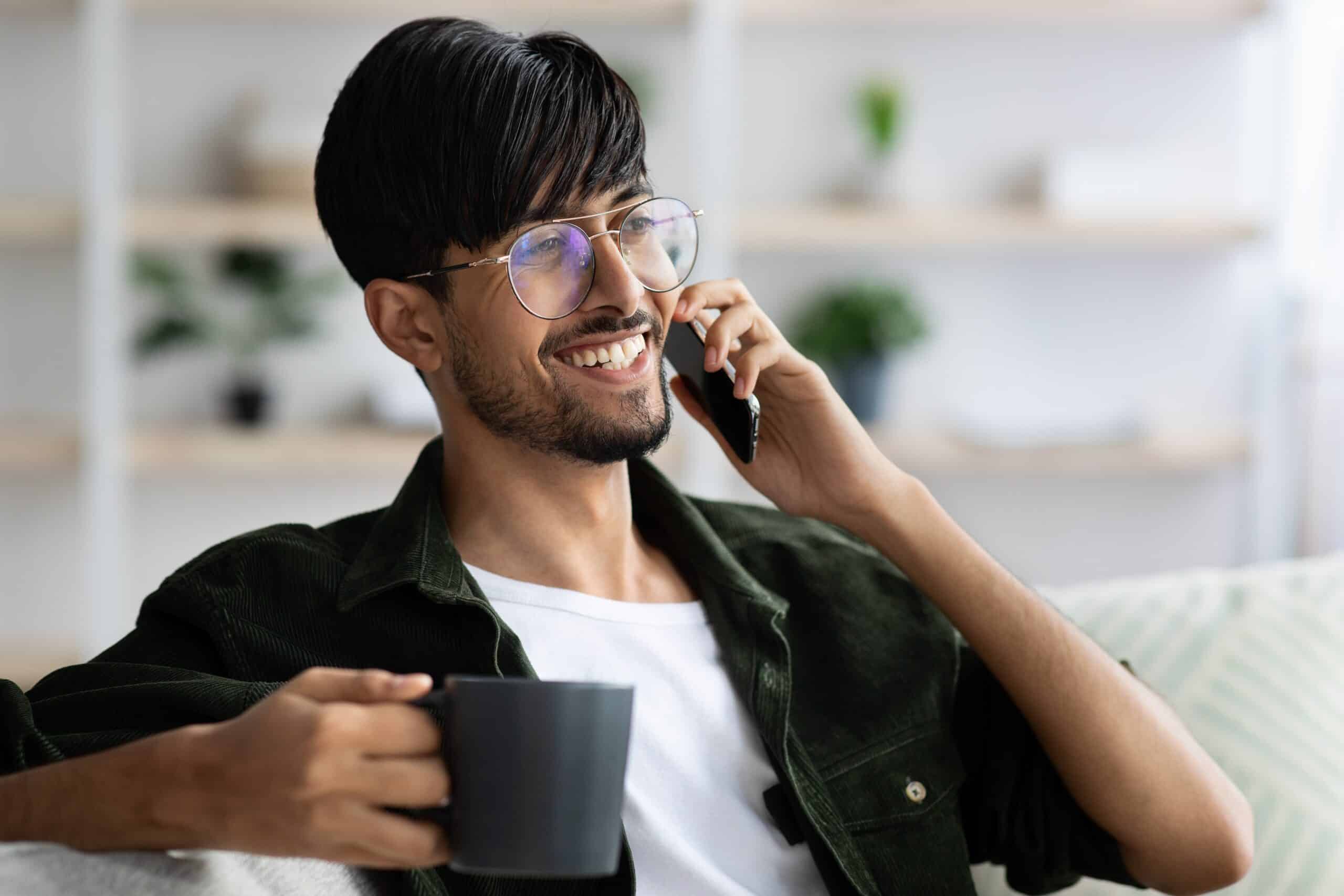 Happy,Young,Indian,Man,In,Glasses,Sitting,On,Sofa,,Talking Smiling man in casual attire holding a coffee cup and talking on the phone, representing personalized financial consultation for Christian schools.