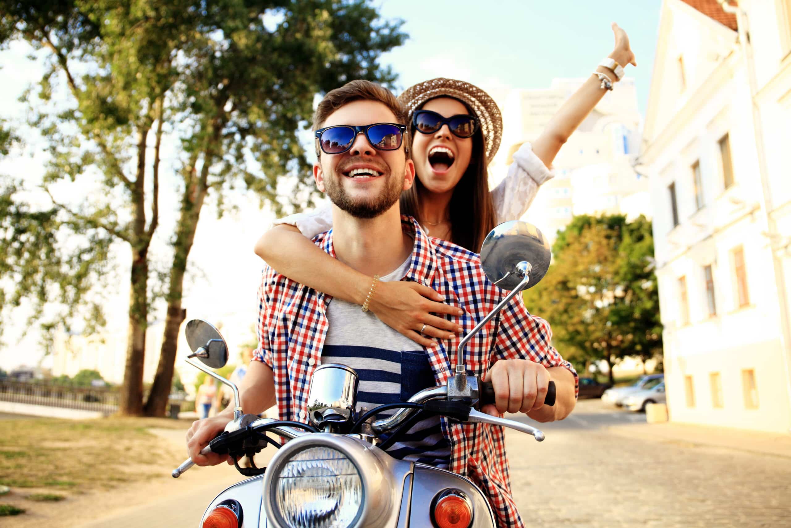 Young couple enjoying a scooter ride, smiling and embracing, in a vibrant outdoor setting, conveying a sense of adventure and joy while travelling.