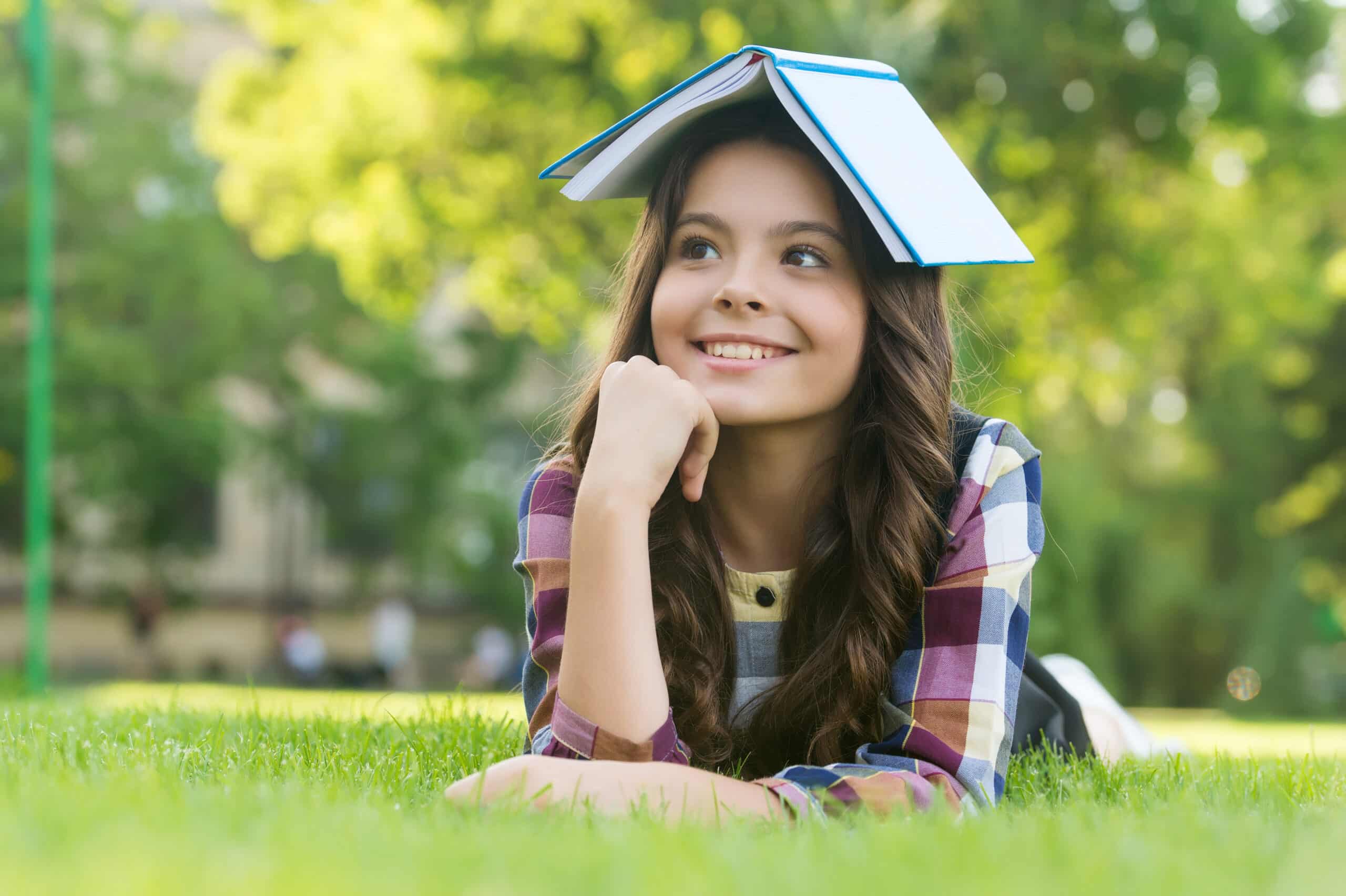Girl lying on grass with a book on her head, smiling and contemplating, representing a positive learning environment for Christian schools.