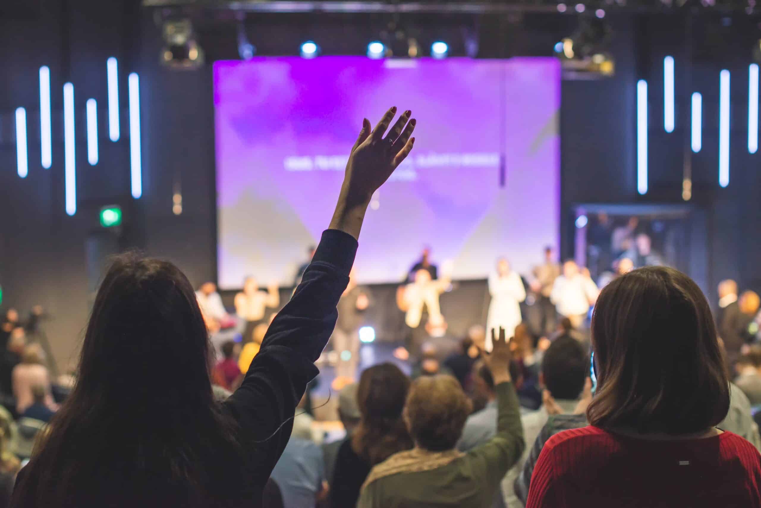 Hands,In,The,Air,Of,People,Who,Praise,God,At Worshippers raising hands during a church service with a vibrant purple screen in the background, reflecting the supportive community environment emphasized by ACS Financial's loan services for churches.