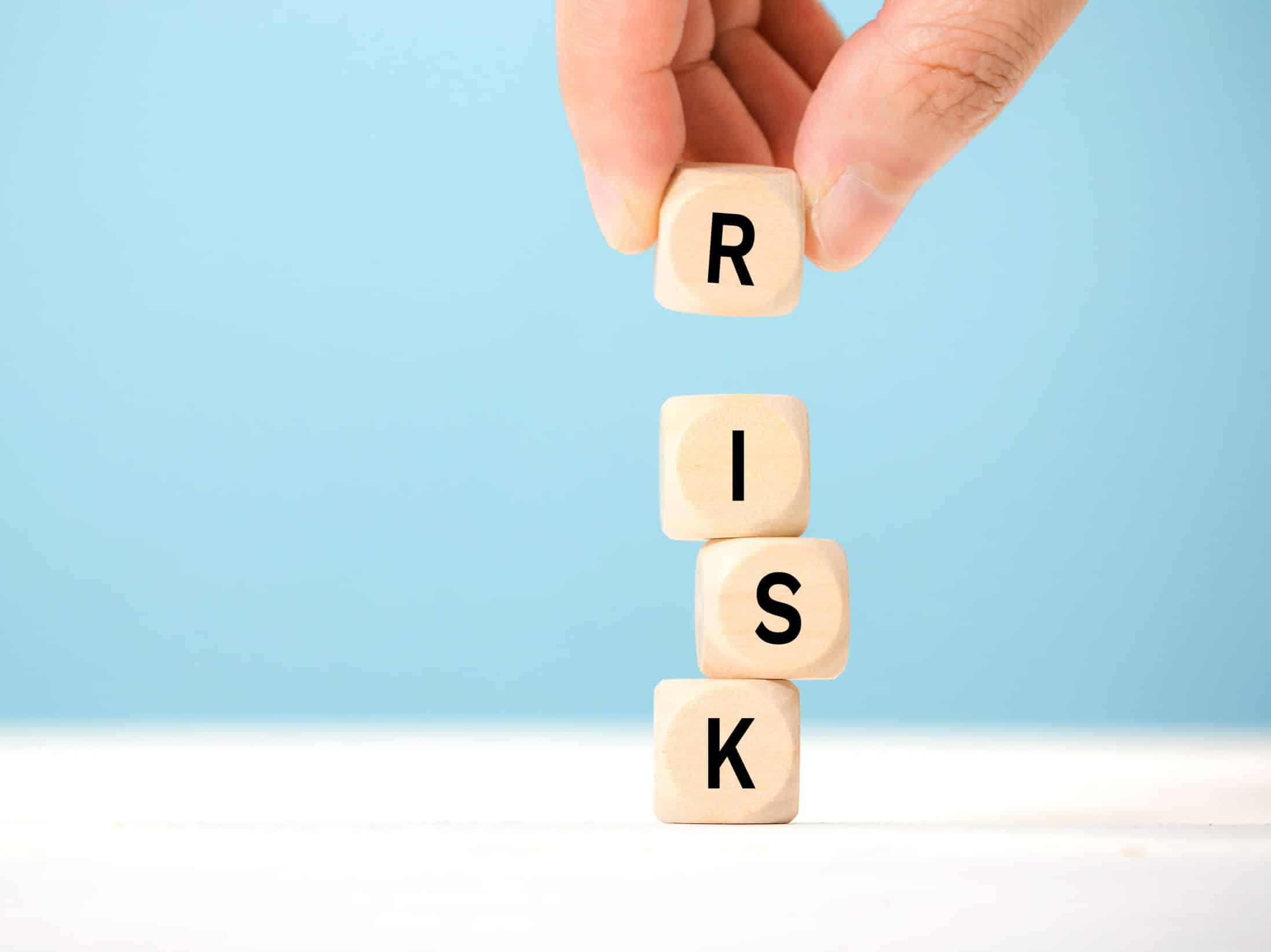 Hand,Hold,Wooden,Cubes,With,Risk,Word.,Risk,Management,Concept. Hand placing wooden blocks spelling "RISK" against a light blue background, symbolizing investment risks in financial services.