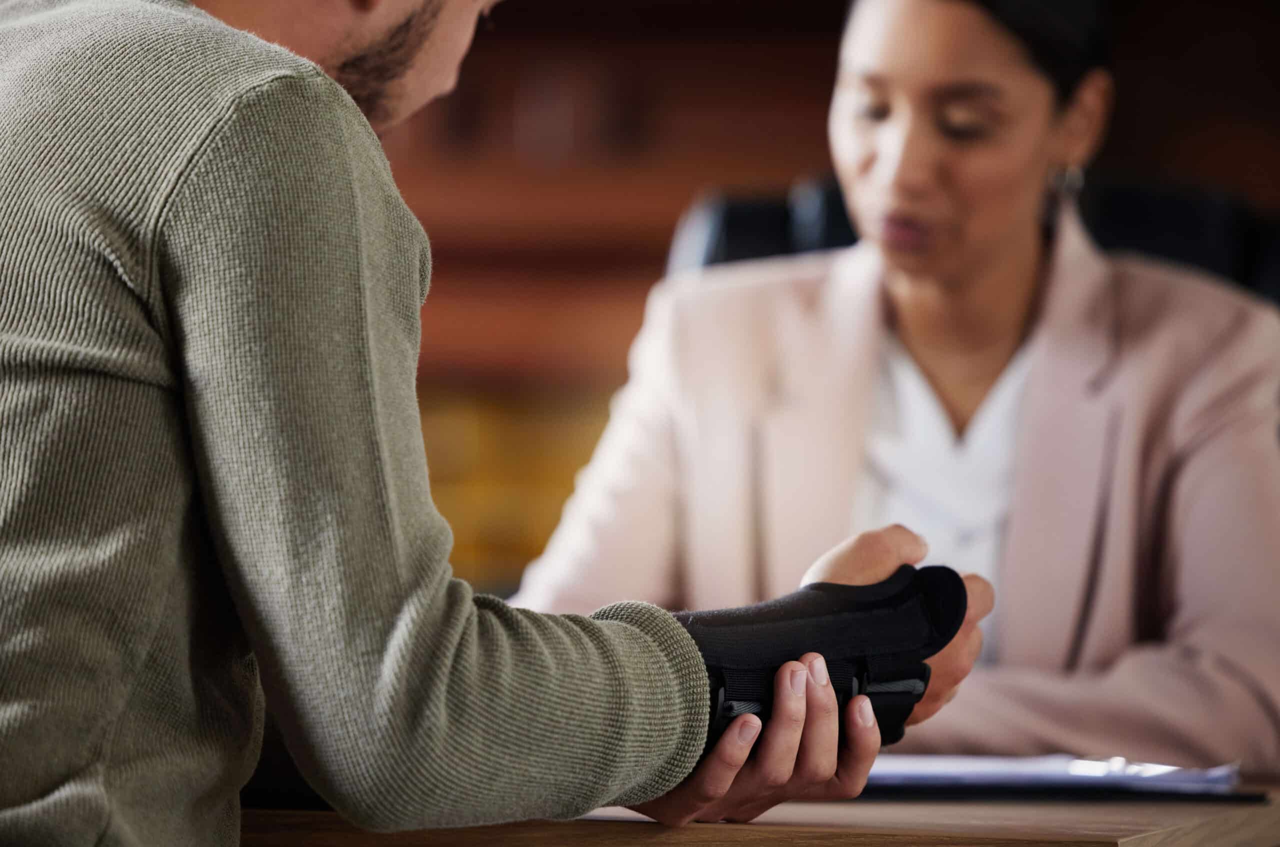 hand-injury-compensation-and-lawyer-people-with-i-2023-03-09-23-14-03-utc (1) Person holding an injured hand in a black brace while discussing compensation options with a lawyer in a professional setting.
