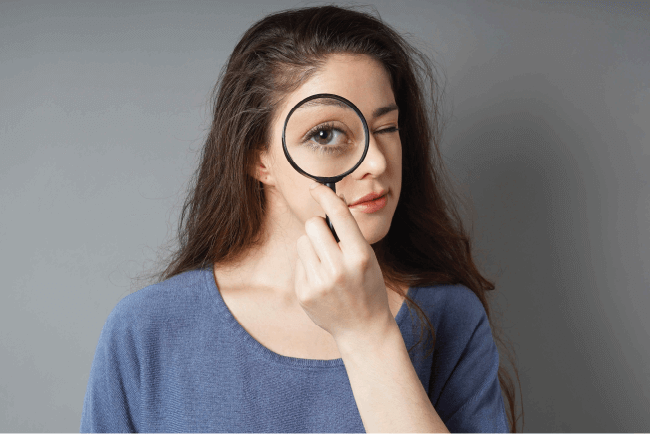 Woman peering through a magnifying glass against a gray backdrop, illustrating cyber risk and fraud inspection.