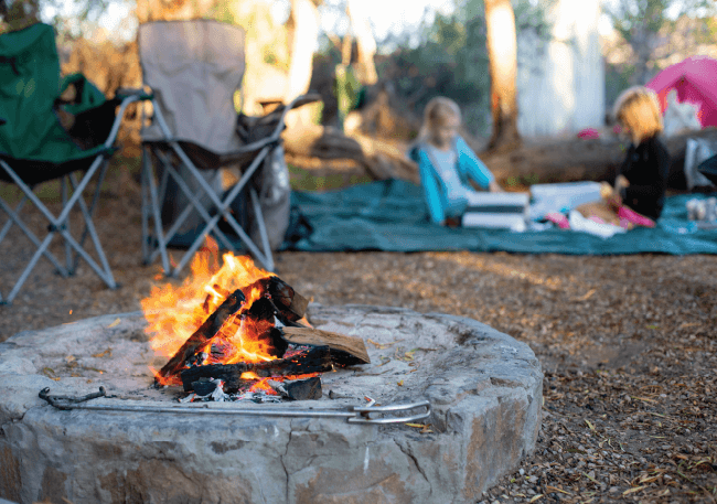 Burning campfire pit at a campsite with youth resting in the background, highlighting church youth camp safety.