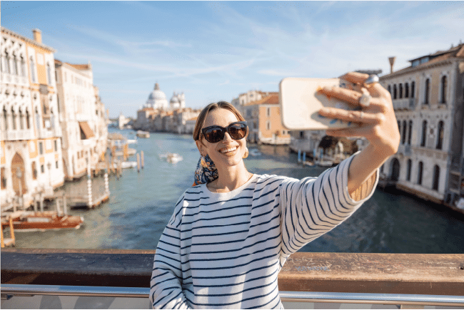 Woman taking a smartphone selfie on a scenic bridge over a grand canal, representing holiday travel insurance plans.