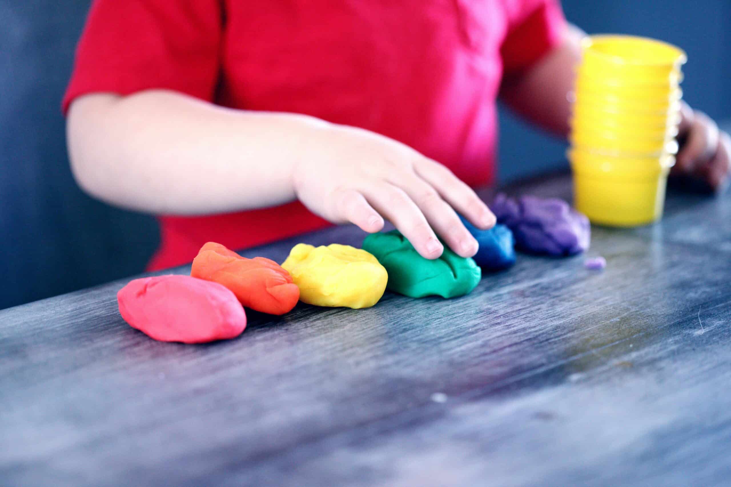 Child's hand reaching for colourful playdough on a table, with yellow cups in the background, illustrating creative play in a childcare setting.