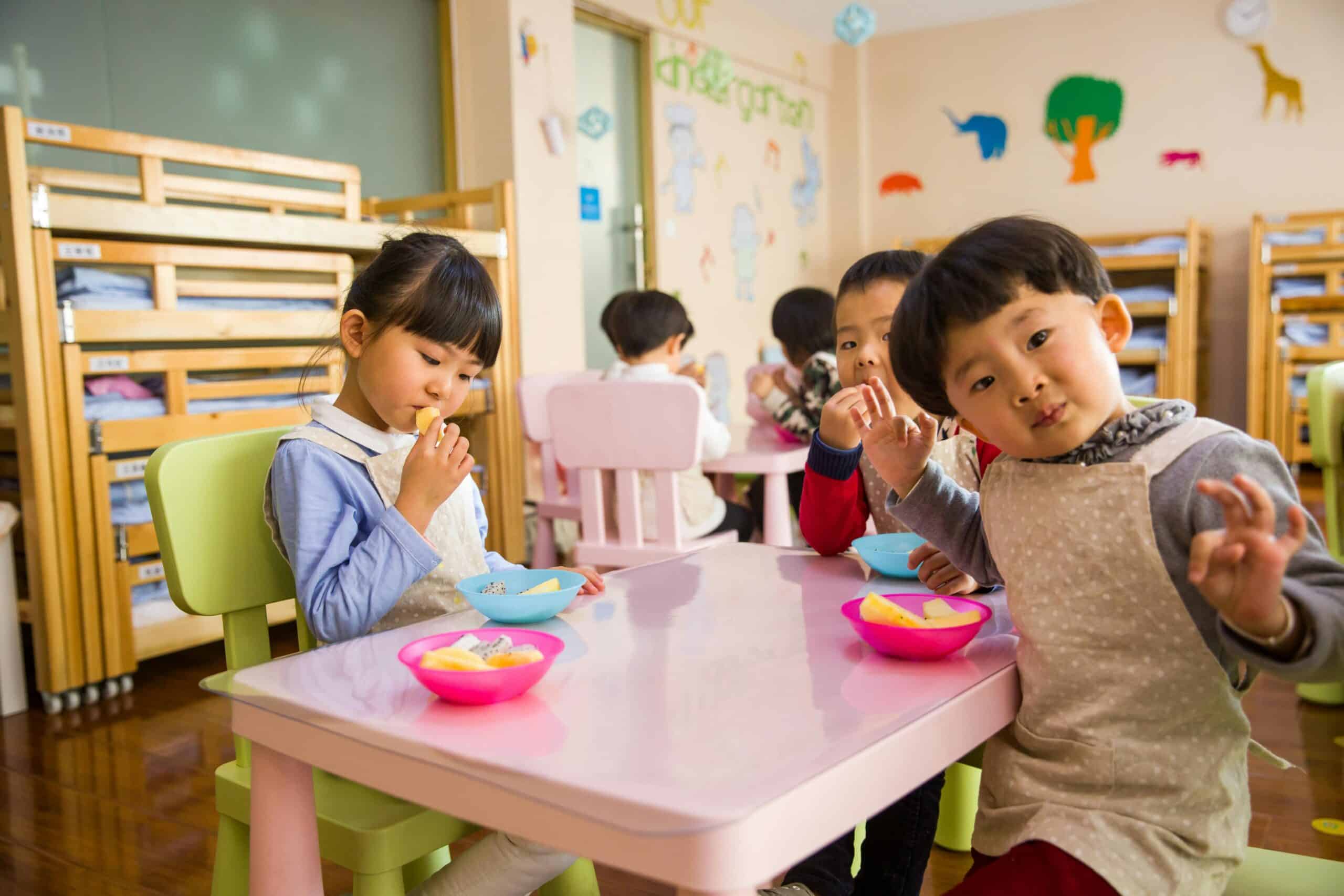 Children enjoying snacks at a daycare table, wearing aprons, with colourful bowls of fruit, in a playful childcare setting.